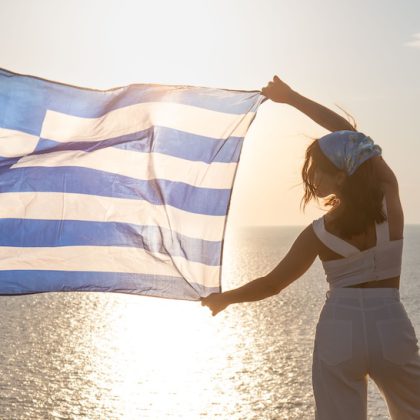 woman with greece flag looking at sunset above the sea Lefkada island lighthouse on background